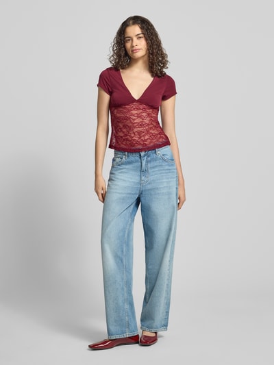 Frauenmodel trägt Damen T-Shirt mit V-Ausschnitt und Spitze von Gina Tricot, Material-Mix mit Stretch-Anteil, Slim-Fit, Rückenlänge 51 cm, Farbkontrast zwischen bordeauxrotem Stoff und transparenter Spitze, blaue Jeans, rote Schuhe, Ganzkörperansicht von vorne.