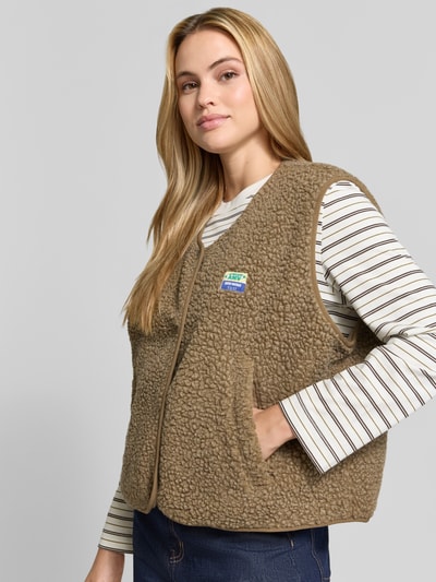 Frauenmodel trägt Regular Fit Weste aus Teddyfell Modell 'Hoktown' von American Vintage, beige, strukturierte Oberfläche, Nahaufnahme Oberkörper. Damen Weste aus Teddyfell, Logo-Patch und Knopfleiste im Fokus, Kontext: Rückenlänge 57 cm, Schulterbreite 43 cm, Brustweite 63 cm.