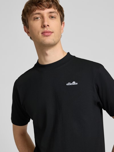 Męski model nosi czarny t-shirt z wyhaftowanym logo, model 'ACCIANO CLASSIC' od Ellesse z bawełny, prosty krój, stójka, nahaufnahme górnej części ciała.