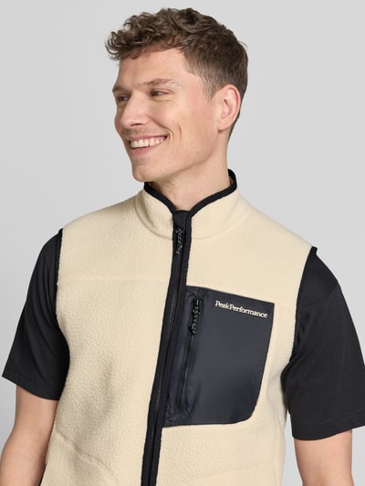 Männermodel trägt Weste mit Brusttasche Modell 'NatureRush' von Peak Performance in Beige mit schwarzer Brusttasche, Nahaufnahme Oberkörper. Fleece, gerader Schnitt, Stehkragen, Zweiwege-Reißverschluss, Logo-Stitching, Reißverschluss-Brusttasche laut Kontext.