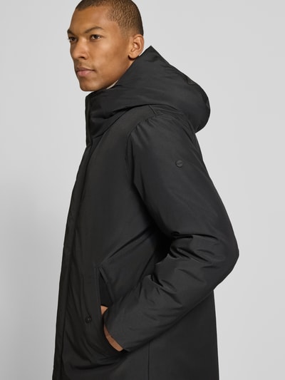 Męski model nosi Jacke mit Label-Patch und Kapuze od khujo w kolorze czarnym, prosty krój, kieszenie z patką, detal z logo, zamek błyskawiczny i rzep, kaptur, Długość tyłu w rozmiarze M: 94 cm, szerokość ramion w rozmiarze M: 49 cm, szerokość klatki piersiowej w rozmiarze M: 55 cm, długość rękawów w rozmiarze M: 68 cm, zdjęcie w półprofilu, widok z przodu.