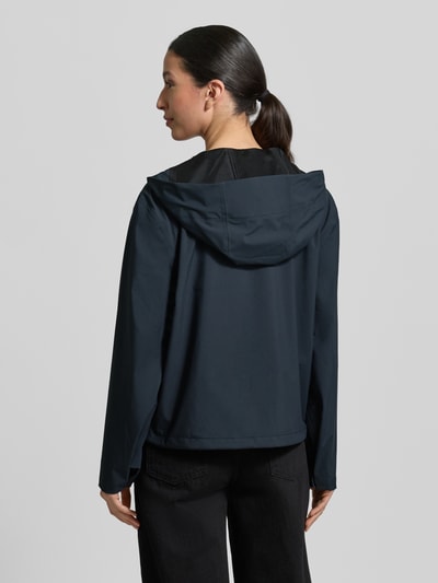 Kobiece modelka ma na sobie kurtkę Regular Fit Jacke mit Kapuze Modell 'Suva' od Rains w kolorze ciemnogranatowym, gładka tekstura, krój regular fit, kaptur z tunelem, zamek błyskawiczny dwustronny, tunel w pasie, długość tyłu 59 cm, szerokość ramion 49 cm, szerokość w rozmiarze S 54 cm, długość rękawów 60 cm, widok całej sylwetki z tyłu.