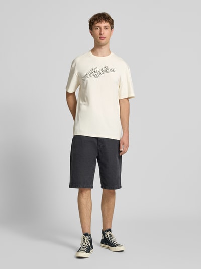 Męski model nosi Oversized T-Shirt mit Logo-Stitching von KARL KANI z czystej bawełny, krój oversized fit, kremowe, czarne logo, okrągły dekolt, długość pleców w rozmiarze M 73 cm, całopostaciowy widok z przodu.