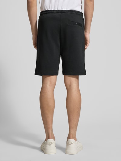 Mężczyzna nosi Shorts mit elastischem Bund von Christian Berg Men w kolorze czarnym, krój regular fit, bawełna z elastanem, szerokość w rozmiarze M 43 cm, długość nogawki 23 cm, kieszenie z kontrastowymi paskami, elastyczny pas z tunelem, widok całej sylwetki od tyłu.