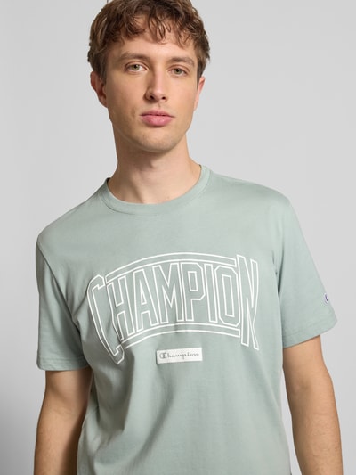 Männermodel trägt T-Shirt mit Logo und Rundhalsausschnitt von CHAMPION aus reiner Baumwolle, Regular-Fit, Label-Stitching, Nahaufnahme Oberkörper.