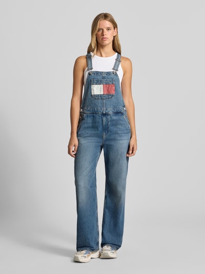 Frauenmodel trägt Relaxed Fit Latzhose aus reiner Baumwolle von Tommy Jeans, mittleres Blau, weiße und rote Details, Ganzkörperansicht von vorne, Damen Latzhose von Tommy Jeans