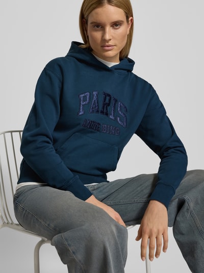 Frauenmodel trägt Regular Fit Hoodie mit Logo-Stitching von Anine Bing in Dunkelblau, Reine Baumwolle, Känguru-Tasche, elastische Abschlüsse, Rückenlänge 58 cm, Ganzkörperansicht von vorne.