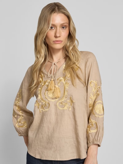 Frauenmodel trägt Leinenbluse mit Motiv-Stitching und Quasten-Details von Emily Van den Bergh in Beige mit gelbem Motiv, Reines Leinen, Loose-Fit, Tunikakragen, Elastik-Ärmelabschlüsse, Rückenlänge 64 cm, Ärmellänge 50 cm, Ganzkörperansicht von vorne.