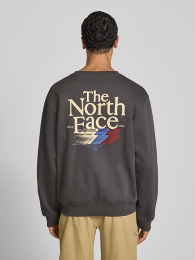 Männermodel draagt Relaxed Fit Sweatshirt aus katoenmix van The North Face met logoprint en ronde hals, herensweatshirt, ruglengte maat M 68 cm, mouwlengte maat M 68 cm, elastische afwerking, donkergrijs met meerkleurig logo, ganzlichaamsaanzicht van achteren