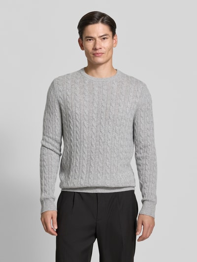 Męski model nosi Slim Fit Strickpullover aus reinem Kaschmir mit Zopfmuster von Christian Berg Men, jasnoszary ścieg warkoczowy, prążkowane wykończenia, okrągły dekolt, krój z długim rękawem, dzianina kaszmir, Ganzkörperansicht von vorne.
