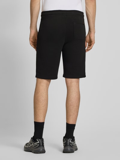 Männermodel trägt Shorts mit Eingrifftaschen und Logo-Print von Alpha Industries in Schwarz, Baumwoll-Mix, Relaxed-Fit, Gesäßtasche, Ganzkörperansicht Rückansicht.