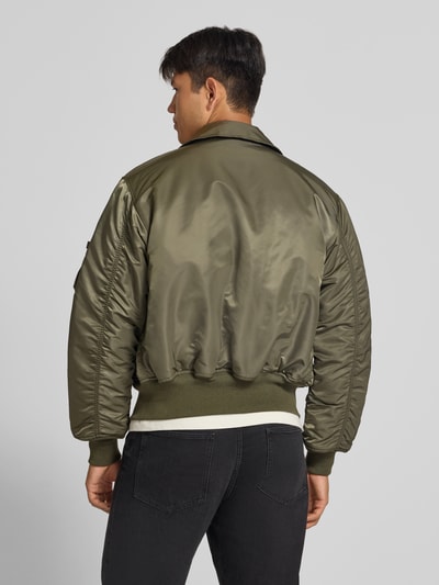 Männermodel trägt Bomberjacke mit seitlichen Pattentaschen und Label-Patch von Alpha Industries in Olivgrün mit gerippten Abschlüssen, Ganzkörperansicht Rückansicht. Regular Fit, Kentkragen, Reißverschluss, Label-Patch und Reißverschlusstasche am Ärmel, Rückenlänge 62 cm, Schulterbreite 50 cm, Brustweite 61 cm, Ärmellänge 67 cm.