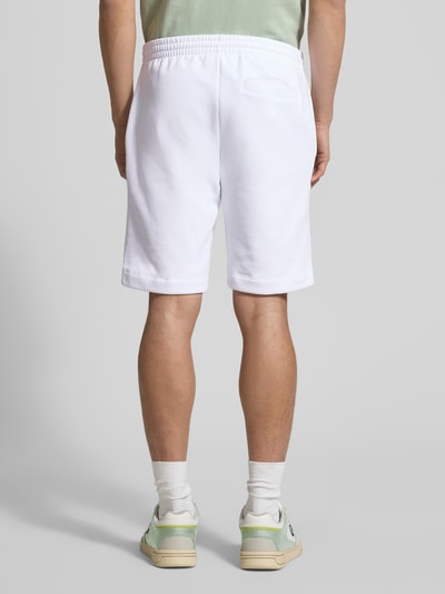 Męski model nosi Regular Fit Sweatshorts aus Baumwoll-Mix od Lacoste, biała tkanina, jednolity kolor, ściągacz w pasie, kieszenie z boku i z tyłu, widok całej sylwetki od tyłu.
