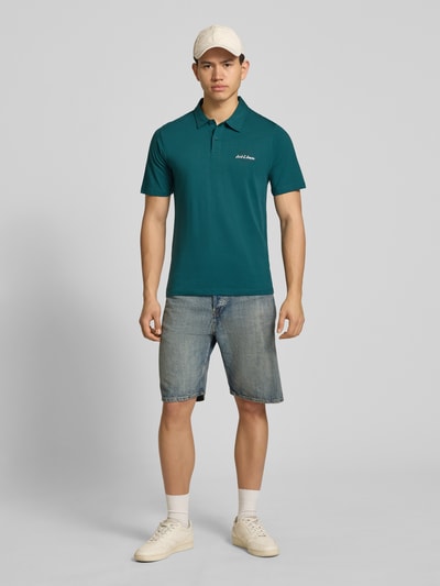 Männermodel trägt poloshirt mit labelprint model 'BRANDON' van Jack & Jones van puur katoen in regular fit, polokraag, korte knoopsluiting, ruglengte bij maat M 71 cm, petrolgroen, ganzkörperansicht van voren.
