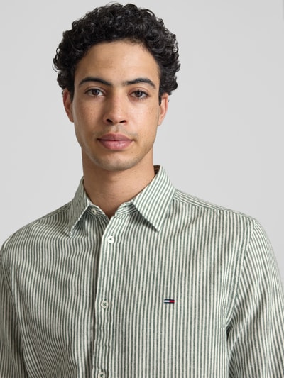 Männermodel trägt Regular Fit Freizeithemd aus Leinen-Baumwoll-Mix von Tommy Jeans, grün-weiß gestreift, Logo, Nahaufnahme Oberkörper.
