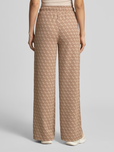 Frauenmodel trägt Sweatpants mit Logo-Muster im Allover-Look von GUESS, Wide Leg, elastischer Bund, beige-brauner Material-Mix, Ganzkörperansicht Rückansicht.