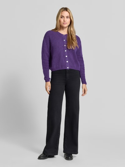 Frauenmodel trägt Relaxed Fit Strickjacke aus Woll-Mix mit Leinen-Anteil von American Vintage in Lila, schwarze Hose, Ganzkörperansicht von vorne. Damen Strickjacke, Woll-Mix, Leinen-Anteil, gerippte Bündchen, Knopfleiste, langärmlig.