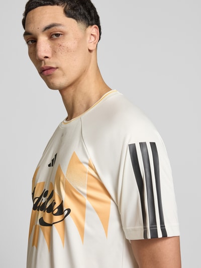 Männermodel trägt T-Shirt mit Logo- und Motivprint von adidas Sportswear in Beige und Gelb, Raglanärmel, Rundhalsausschnitt, Lockerer-Schnitt, Nahaufnahme Oberkörper.