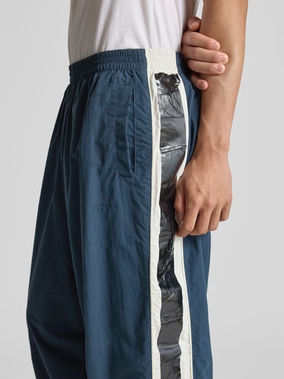 Männermodel trägt Trackpants mit elastischem Bund von VETEMENTS in Blau mit weißen und schwarzen Galonstreifen, seitlichen Eingrifftaschen, Mesh-Futter, Nahaufnahme Oberkörper.