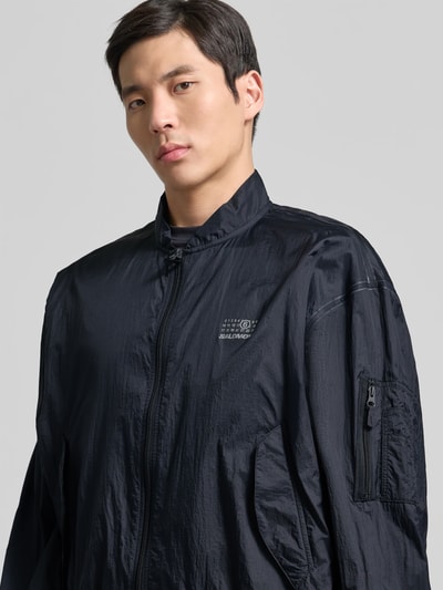 Männermodel trägt Relaxed Fit Jacke mit Logo-Print von MM6 Maison Margiela in Schwarz, Reißverschluss, elastische Abschlüsse, Nahaufnahme Oberkörper.