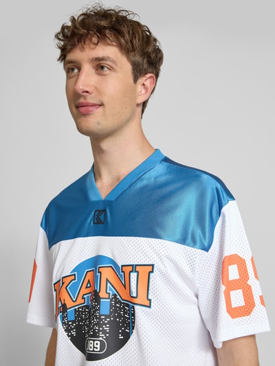 Männermodel trägt Trikot-Shirt mit Logo- und Motiv-Prints Modell 'OG Sport College' von KARL KANI in Blau, Weiß und Orange, Nahaufnahme Oberkörper, Boxy-Fit, V-Ausschnitt, Rückenlänge bei Größe M: 64 cm.