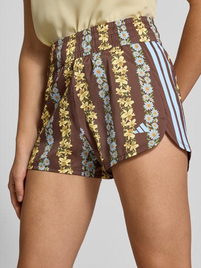 Frauenmodel trägt Regular Fit Sportshorts mit Allover-Muster Modell 'FARM SHORTS' von adidas, braun mit gelbem und blauem Muster, Damen Sportshorts von adidas Sportswear, Nahaufnahme Oberkörper.