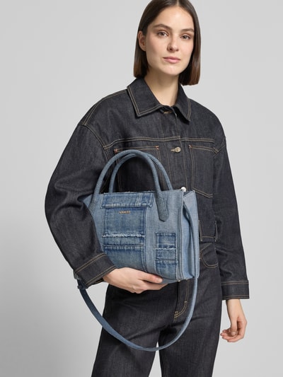 Frauenmodel trägt Tote Bag mit Viskose-Anteil Modell 'Jodi' von Guess, Denim-Optik, Material-Mix, Logo-Detail, Hauptfach mit Reißverschluss, Schulterriemen und Tragehenkel, 34 cm breit, 24 cm lang, 11 cm tief, Schulterriemen 110 cm, Ganzkörperansicht von vorne.