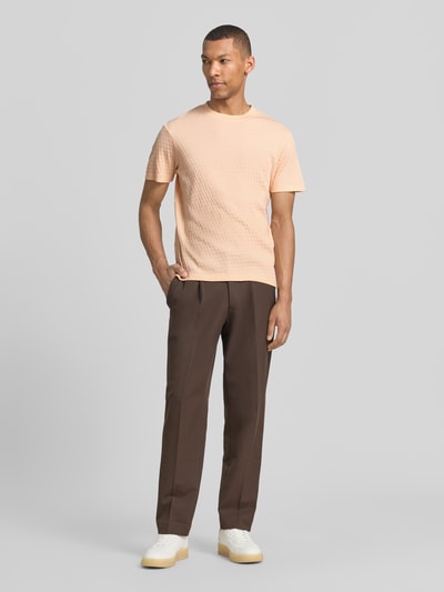 Männermodel trägt T-Shirt mit Strukturmuster Modell 'Raulos' von JOOP! Collection in Hellorange, Reine Baumwolle, Modern Fit, Rundhalsausschnitt, Label-Detail, kombiniert mit brauner Hose, Ganzkörperansicht von vorne.
