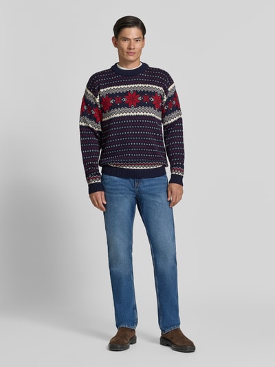 Männermodel trägt Regular Fit Wollpullover in Strick-Optik Modell 'Egersund' von Dale of Norway in Dunkelblau, Rot und Weiß, Ganzkörperansicht von vorne. Herren Wollpullover aus reiner Wolle, Rückenlänge 64 cm, Ärmellänge 58 cm.