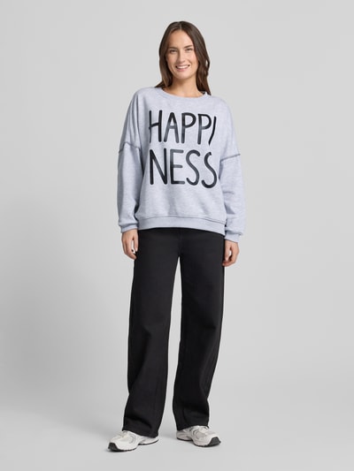 Frauenmodel trägt Sweatshirt mit Statement-Print von miss goodlife in Hellgrau, Reine Baumwolle, Lockere Schnitt, Gerippte-Abschlüsse, Rundhalsausschnitt, schwarze Hose, helle Sneaker, Ganzkörperansicht von vorne.