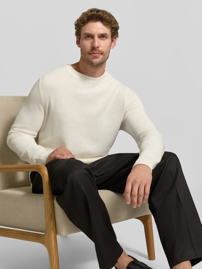 Męski model nosi Regular Fit Pullover aus reiner Baumwolle Modell 'Lalex' od BOSS, sweter z bawełny w kolorze jasnym, ciemne spodnie, tekstura dzianiny, zdjęcie: siedzący, ujęcie całej sylwetki z przodu.