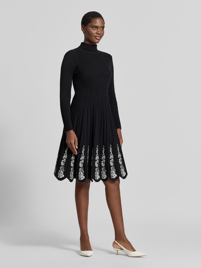 Modelka kobieca nosi knielanges Strickkleid aus Viskose-Mix von Apricot, czarny kolor, Efekt prążkowania, taliowany krój, Stójka, krój z długim rękawem, spódnica linia a z plisowaniem, hafty kwiatowe, Długość do kolan, Długość tyłu w rozmiarze S: 99 cm, Szerokość w rozmiarze S: 34 cm, Długość rękawów w rozmiarze S: 60 cm, widok całej sylwetki z przodu