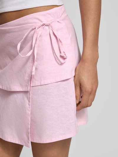 Frauenmodel trägt Minirock mit Leinen-Baumwoll-Anteil Modell 'Sina' von JJXX in Rosa, Zierschleife, Wickel-Optik, Nahaufnahme Oberkörper.