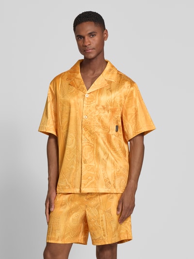Mężczyzna model nosi Relaxed Fit Freizeithemd mit Reverskragen Modell 'Resort' od COLUCCI, elastyczna mieszanka materiałów, krój relaxed fit, kołnierz z połami, krótki rękaw, brustasza, listwa guzikowa, naszywka z logo, długość tyłu w rozmiarze M 70 cm, szerokość w rozmiarze M 56 cm, długość rękawów w rozmiarze M 26 cm, żółtopomarańczowe barwy z delikatnym wzorem paisley, widoczna gładka tekstura, cała sylwetka z przodu