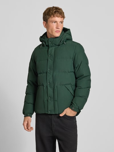 Männermodel trägt grüne Jacke mit Kapuze Modell 'CLASSIC EVEREST' von Superdry, Reiß- und Druckknopfverschluss, Ganzkörperansicht von vorne.