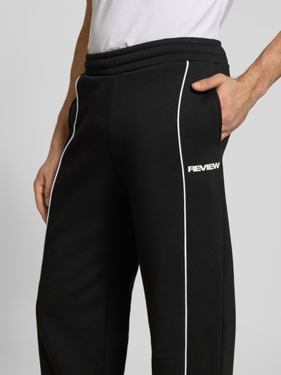 Męski model nosi spodnie dresowe z elastycznym pasem i logo od REVIEW, czarne z białymi lamówkami, krój relaxed fit, bawełna, długość wewnętrzna nogawki w rozmiarze M: 78 cm, detaliczny widok na biodra, ujęcie z przodu