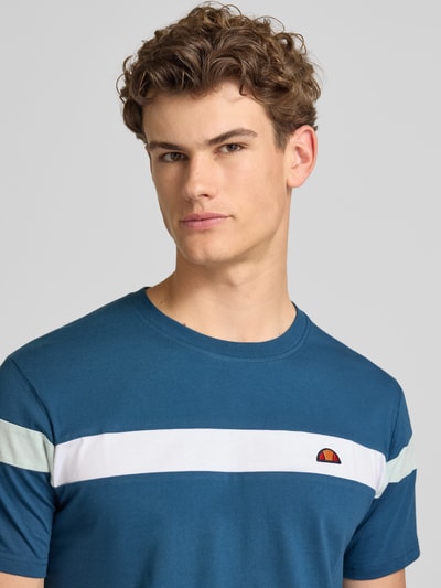 Męski model nosi Regular Fit T-Shirt mit Logo-Detail Modell 'Caserio' od Ellesse z czystej bawełny, okrągły dekolt, krótki rękaw, długość tyłu 68 cm w rozmiarze M, kolor niebieski z białym paskiem, detal z logo, zbliżenie na górną część ciała.