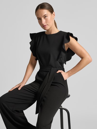 Frauenmodel trägt Jumpsuit mit Rundhalsausschnitt und Bindegürtel von Apricot in Schwarz, glatte Textur, Regular Fit, Nahaufnahme Oberkörper. Material-Mix mit Stretch-Anteil, detailreiche Rüschenärmel sichtbar.