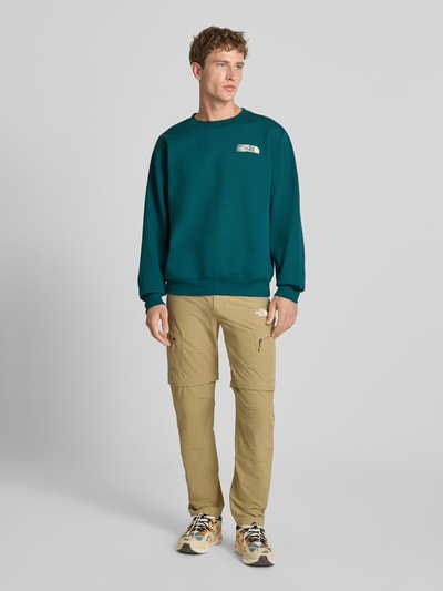 Männermodel trägt Relaxed Fit Sweatshirt aus Baumwoll-Mix von The North Face in Dunkelgrün mit Logo-Print, Ganzkörperansicht von vorne. Baumwoll-Mix, elastische Abschlüsse, Rundhalsausschnitt, Rückenlänge und Ärmellänge je 68 cm bei Größe M.