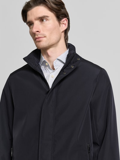 Männermodel trägt Trenchcoat mit Stehkragen von ARMANI EXCHANGE in Dunkelblau, glatte Oberfläche, verdeckter Zipper, Nahaufnahme Oberkörper. Gerader Schnitt, Reißverschlusstaschen, Größe M: Rückenlänge 88 cm, Schulter 49 cm, Brust 58 cm, Ärmel 66 cm.