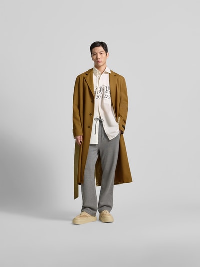 Männermodel trägt Mantel aus reiner Schurwolle von MM6 Maison Margiela in Braun mit Reverskragen und Taillengürtel, Ganzkörperansicht von vorne.
