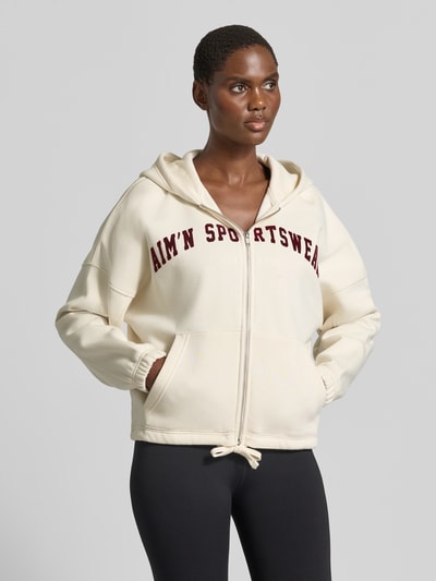Frauenmodel trägt Sweatjacke mit Logo-Schriftzug von AIMN in Creme mit Kängurutasche und Kapuze, Regular Fit, Ganzkörperansicht von vorne.