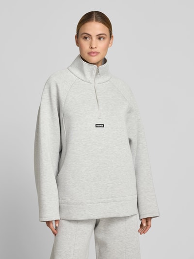 Frauenmodel trägt Sweatshirt aus Viskose-Mix von ROTATE in Hellgrau mit Logo-Patch, Stehkragen, Reißverschluss, taillierter Passform, Struktur glatt, Rückenlänge bei Größe S 69 cm, Ganzkörperansicht von vorne.