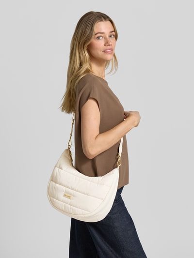 Frauenmodel trägt Halfmoon Bag mit Label-Detail Modell 'Lifetime Buddy M' von Hey Marly in Creme, wattierte Struktur, goldene Details, abgerundete Form, Damen Schultertasche mit Reißverschluss, verstellbarem Gurt und Innenfach, Ganzkörperansicht von vorne.