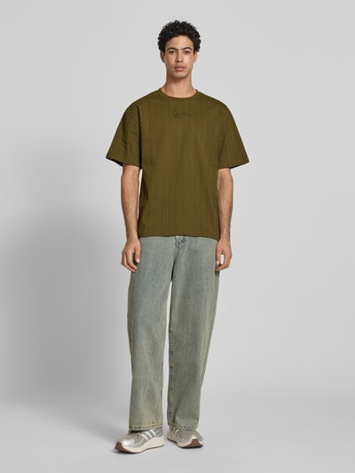 Männermodel trägt Relaxed Fit T-Shirt mit Logo-Stitching von KARL KANI in Oliv, Reine Baumwolle, Rundhalsausschnitt, Rückenlänge 71 cm, Ganzkörperansicht von vorne.
