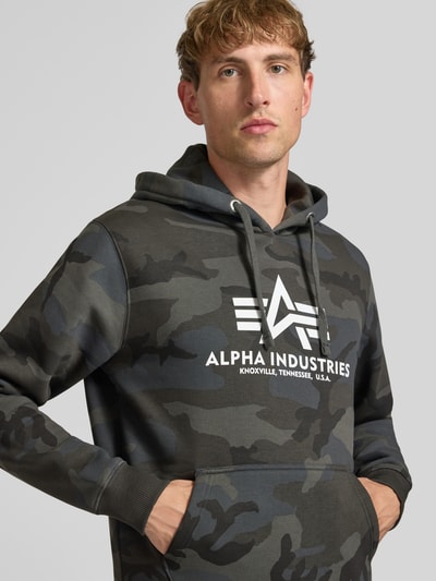 Männermodel trägt Regular Fit Hoodie aus Baumwoll-Mix mit Logo-Print von Alpha Industries, Camouflage-Muster, Nahaufnahme Oberkörper.