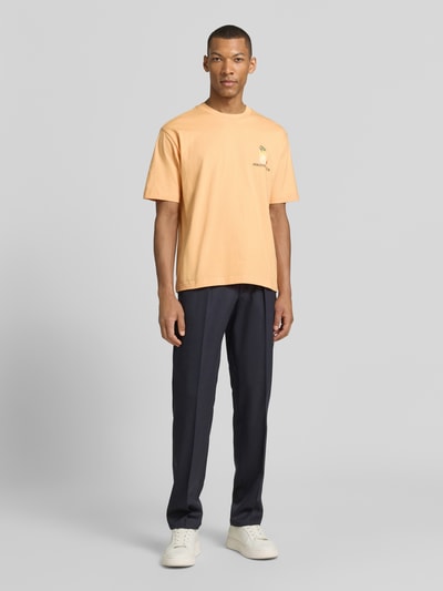 Männermodel trägt T-Shirt mit rückseitigem Motiv-Print von MCNEAL in oranje, rechte pasvorm, ronde hals, biologisch katoen, voorkant bedrukking, volledige lichaamsvoorzijde