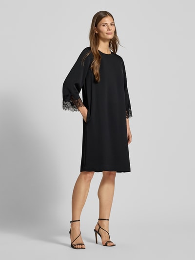 Kobiece modelka nosi sukienkę Knielanges Kleid mit Rundhalsausschnitt Modell 'Dancia' von JOOP! Collection, czarny kolor, krój relaxed fit, okrągły dekolt, długość do kolan, rękaw o dł. 3/4 z obszyciem koronką, długość tyłu 100 cm w rozmiarze 36, szerokość klatki piersiowej 49 cm, długość rękawa 65 cm, gładka tekstura, całkowity widok z przodu.