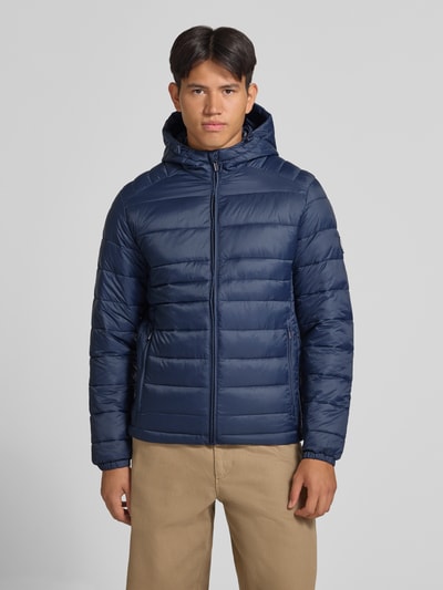 Męski model nosi kurtkę Regular Fit Jacke in Stepp-Optik Model 'KINGSLEY' od JACK & JONES, kolor granatowy, z efektem pikowania, kaptur, zamek błyskawiczny, wpuszczane kieszenie, długość tyłu w rozmiarze M: 70 cm, szerokość ramion w rozmiarze M: 45 cm, szerokość klatki piersiowej w rozmiarze M: 52 cm, długość rękawów w rozmiarze M: 67 cm, zdjęcie całej sylwetki z przodu.