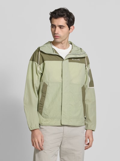 Männermodel trägt Windbreaker mit verdecktem Reißverschluss von Columbia in Grün, Regular-Fit, Kapuze mit Tunnelzug, Logo, Nahaufnahme Oberkörper. Rückenlänge bei Größe M: 70 cm, Schulterbreite: 64 cm, Brustweite: 57 cm, Ärmellänge: 56 cm.